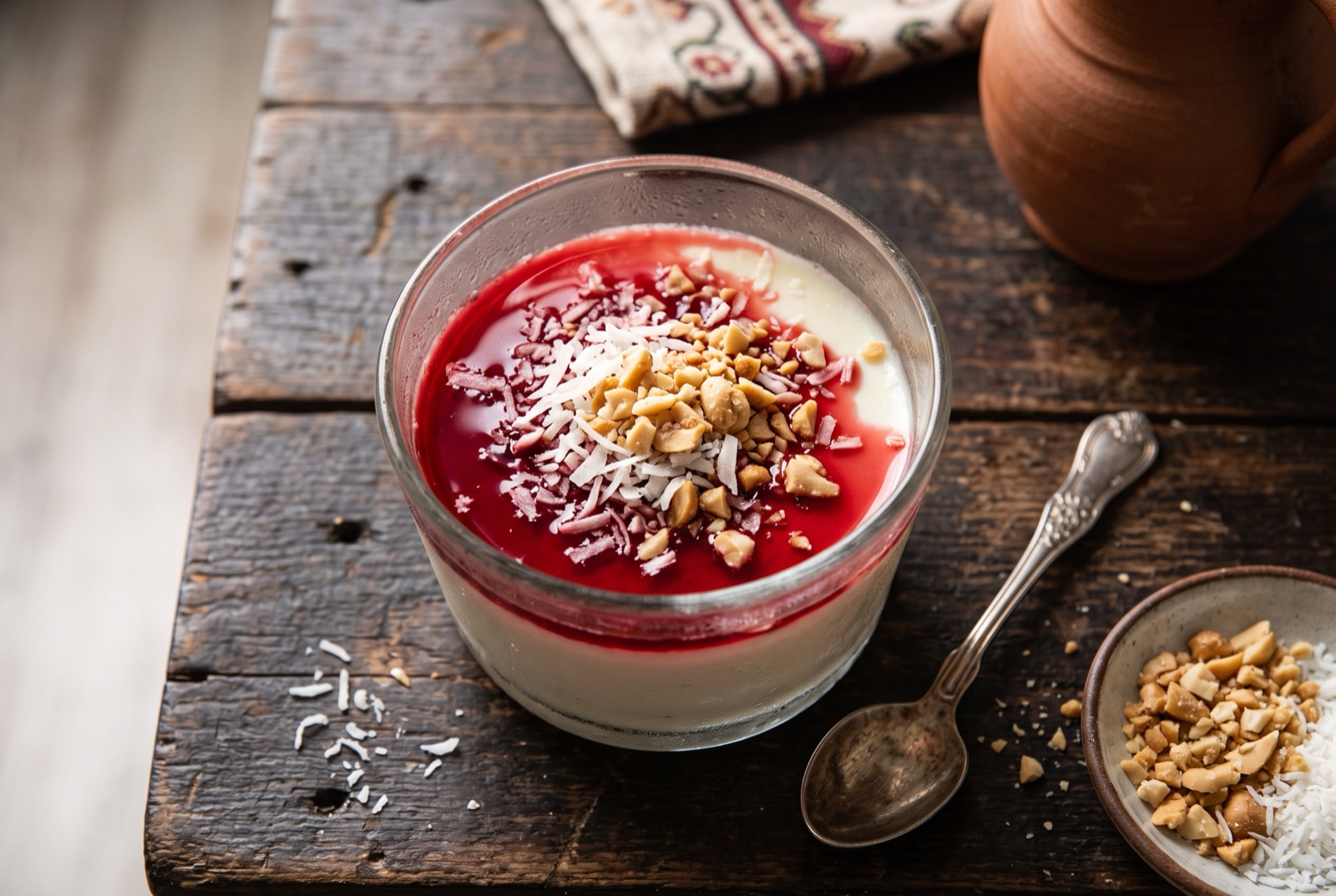Glass of pale set milk pudding under bright red syrup, topped with shredded coconut and crushed peanuts