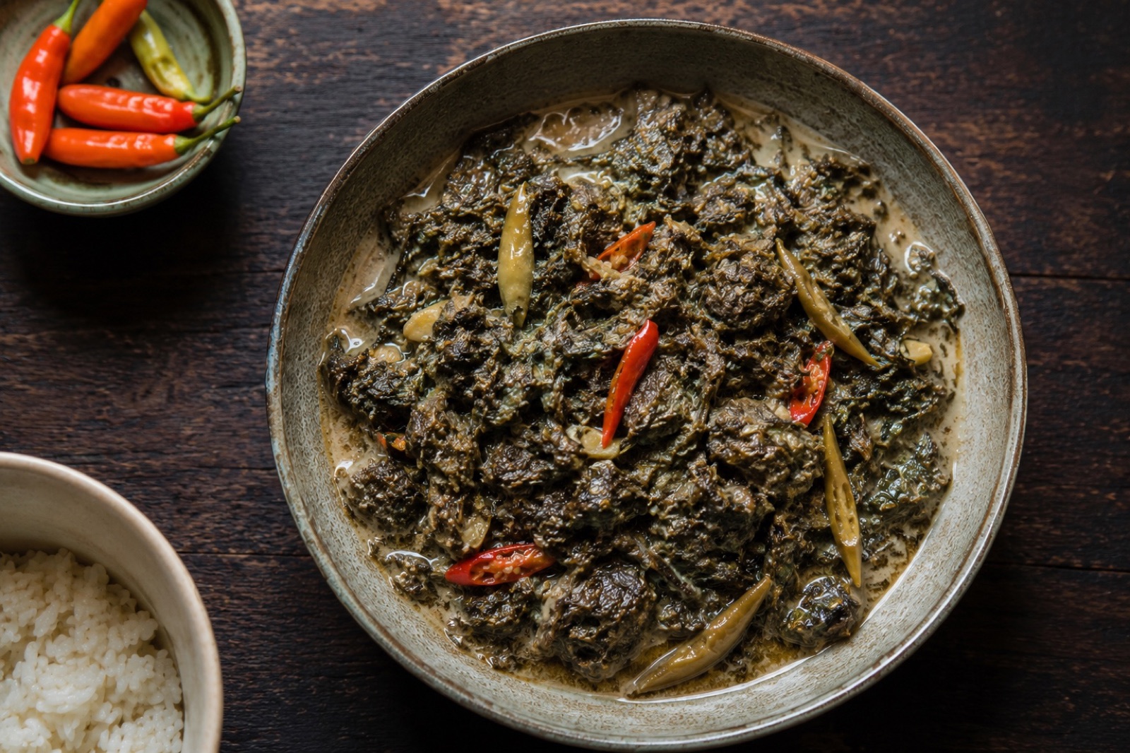 Dark laing of collapsed taro leaves in coconut milk with whole green chiles and coconut oil pooling on the surface